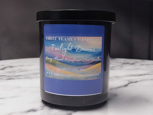 Twilight Dreams scented candle by Three Flames Candle Co, featuring honey, almond, moonflower, chai, and vanilla fragrances. 11 oz black jar with colorful label showing sunset hues, displayed on marble surface.