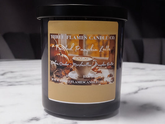 Black candle jar with Twisted Pumpkin Latte label featuring autumn-themed imagery, including a coffee cup and pumpkins. The label showcases warm, inviting colors reminiscent of fall, set against a marble surface.